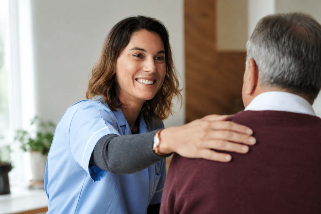A smiling female caregiver in blue scrubs gently places her hand on an older man's shoulder while speaking with him in a bright indoor setting.